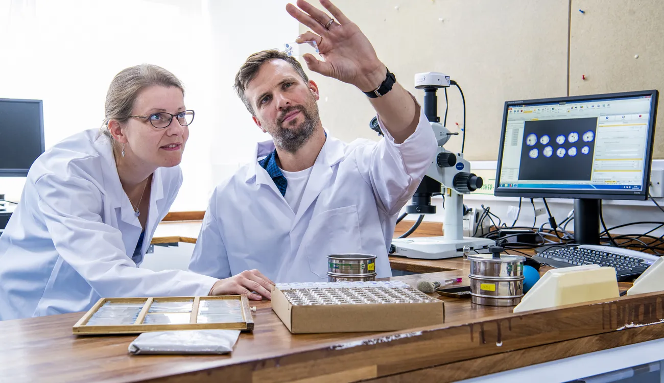 Two researchers wearing white lab coats, work together to prepare samples in laboratory with a computer.