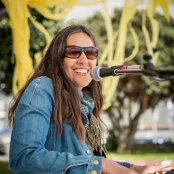 Emily Macklow is sitting playing a piano and has a microphone in front of her face. She is smiling and looking at the camera.