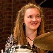 Holly Barnett sitting at her drum kit. She is smiling looking up to the camera.