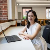 Lin sitting at a desk in an empty classroom. She has a laptop on the desk in front of her, and she's smiling for the camera.