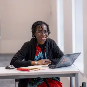 Ogene sat at a desk with a laptop and notebook in front of her. She's looking at the camera and smiling.