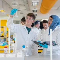 Two students wearing lab coats and protective glasses conduct an experiment. One holds up a glass beaker, while the other takes notes. They are in a laboratory, surrounded by a variety of chemistry equipment.