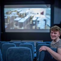A student leaning over the back of his seat in a cinema as a film plays in the background, smiling at the camera.