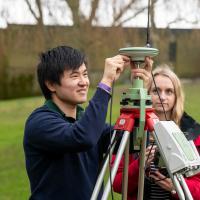 Two students conduct an experiment outdoors using a large piece of survey equipment on a tripod.
