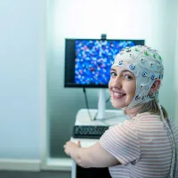 A student sits in front of a computer and turns to smile towards the camera. She is wearing a head cap that is connected to electrodes to monitor brain activity.