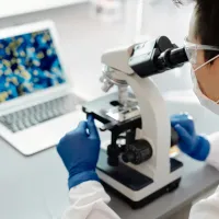 Student in a lab coat is looking into a microscope and showing results on the laptop infront of her