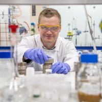A chemistry researcher in goggles, gloves and white coat works with equipment in the lab