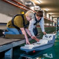 A student crouches down to place a model boat into the water of the university towing tank, a researcher at her side