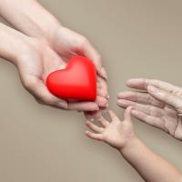 A pair of hands holding a heart-shaped object reach out, while a baby's hand and an old person’s hand reach up towards it. 