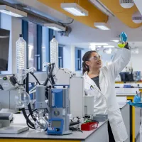 Two students in white coats in the newly refurbished chemistry labs with distillation glass ware set up. They are wearing safety glasses and looking up at a flask containing a yellow liquid. 