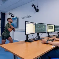 A man performs a lunging squat with motion capture markers, while another man monitors data on screens in a lab.