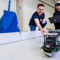 Two students testing a small mobile robot within a bounded arena in university robotics lab.