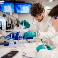 Students wearing safety goggles and gloves conduct a laboratory experiment using pipettes and glassware.