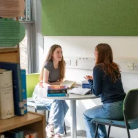 Two students sitting at a table in a library, studying and discussing books together.