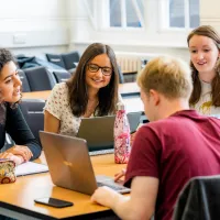 Five students gathered around a table using laptops and discussing work together.